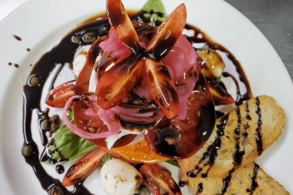 A colorful salad topped with tomatoes, pickled onions, and drizzled with balsamic glaze, served with toasted bread.