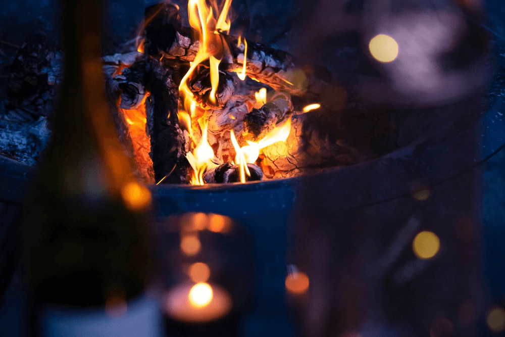 A glowing fire burns in a fire pit, with a blurred bottle and candles in the foreground.