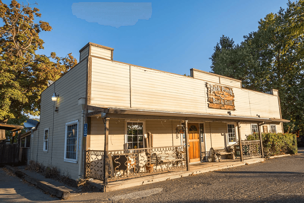 A rustic bar and grill with a wooden sign and landscaped entrance on a sunny day.