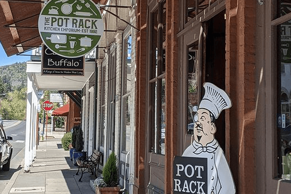 A chef sign stands outside "The Pot Rack," a kitchen emporium with a green sign, on a sunny street.