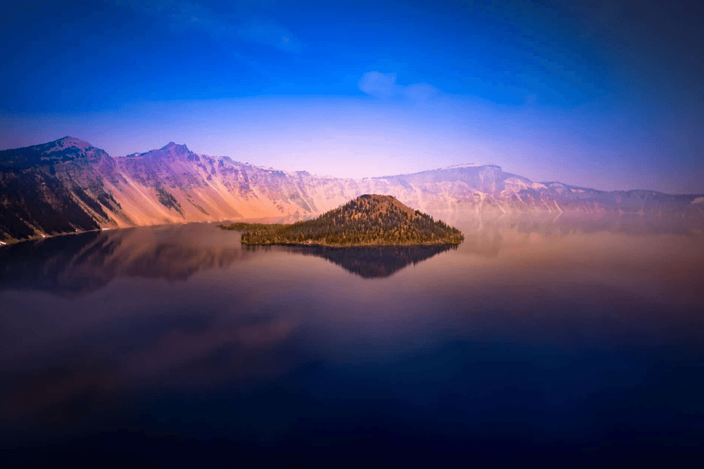 A tranquil lake surrounded by mountains with a small island in the center, reflecting the colorful sky.