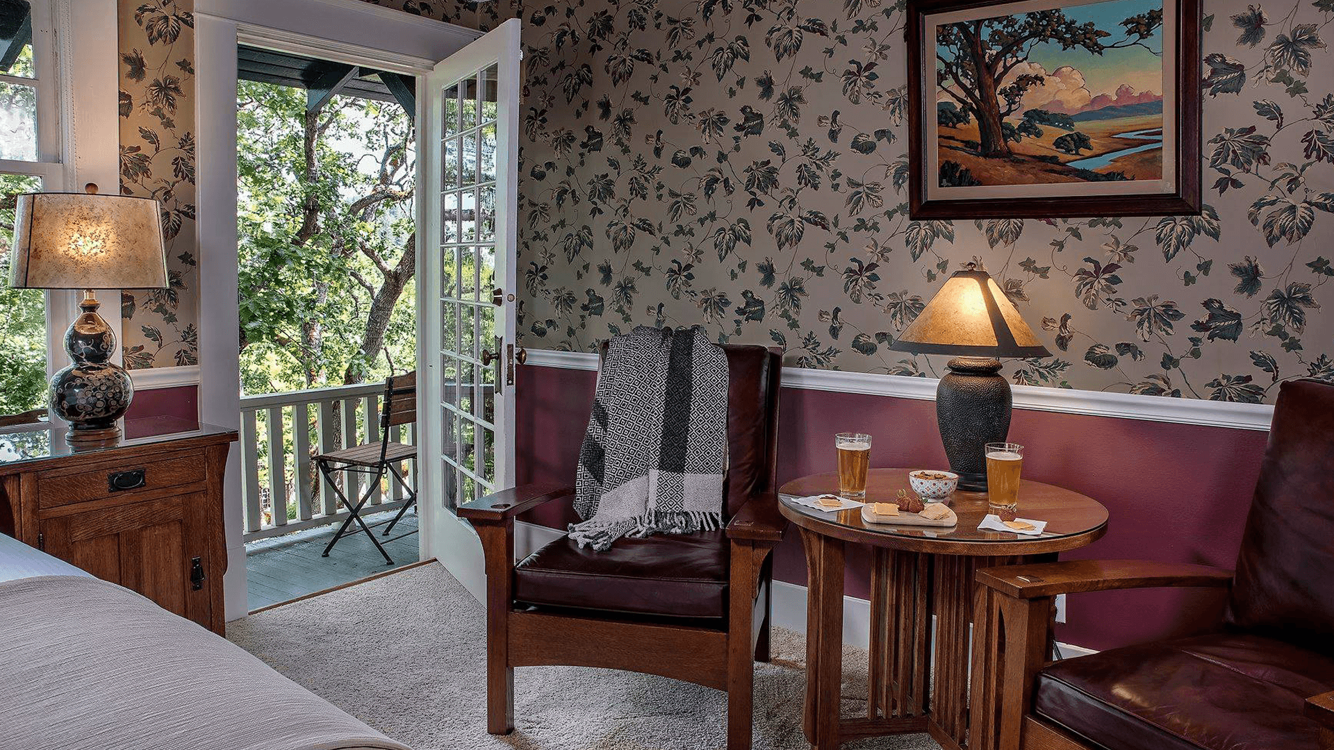 A cozy room featuring a patterned wall, two chairs, a side table with drinks, and a view of a balcony through an open door.
