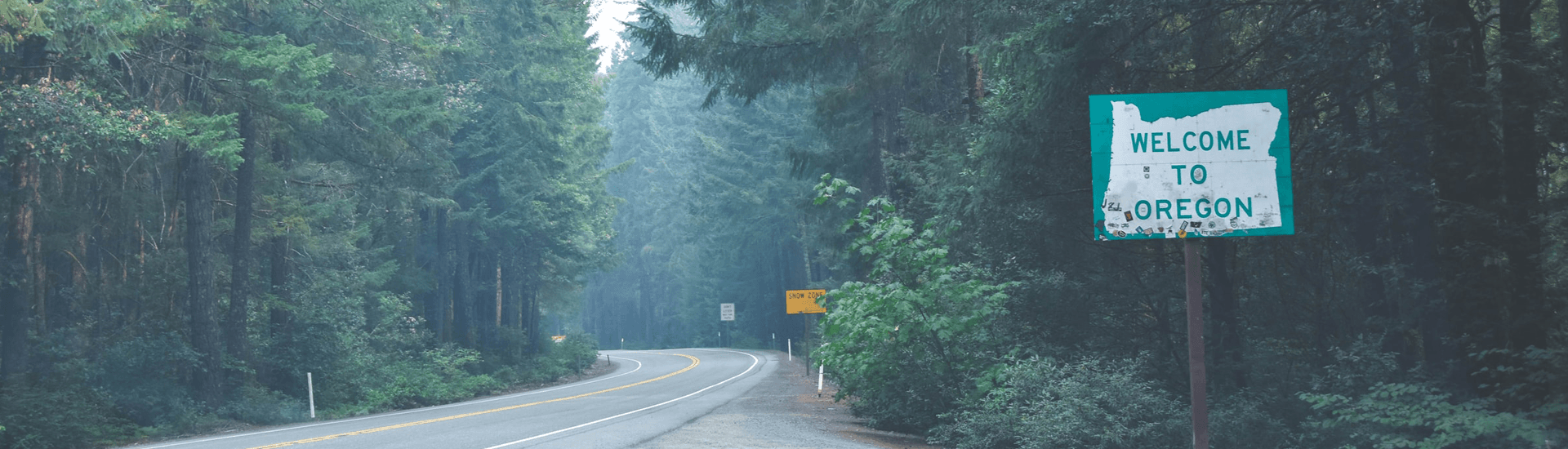 A "Welcome to Oregon" sign alongside a winding road surrounded by tall trees.